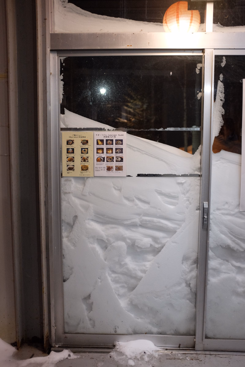 A glass door covered in thick snow, with a menu visible inside that reads "メニュー".