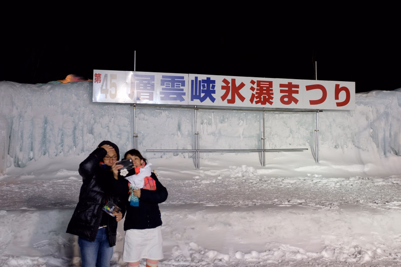 Two people stand in front of a large sign that reads "第45雪映氷まつり" during a winter ice festival.