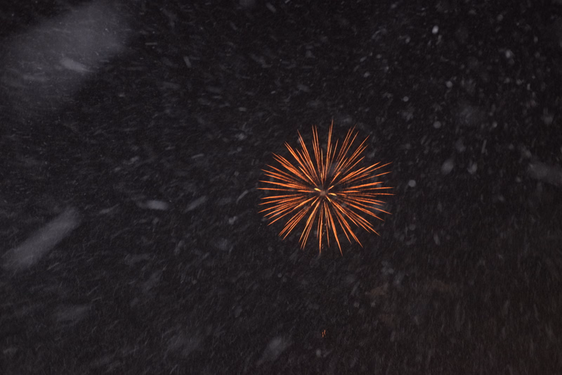 A bright burst of fireworks illuminates the dark night sky while snow falls around it.