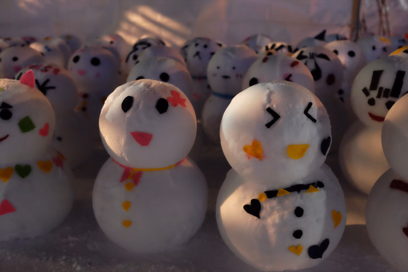A collection of small, decorated snowmen are lined up on a snowy surface, some with colorful accents.