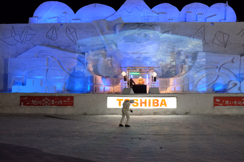 A person walks past a large illuminated display featuring the text "TOSHIBA" on a beach at night.