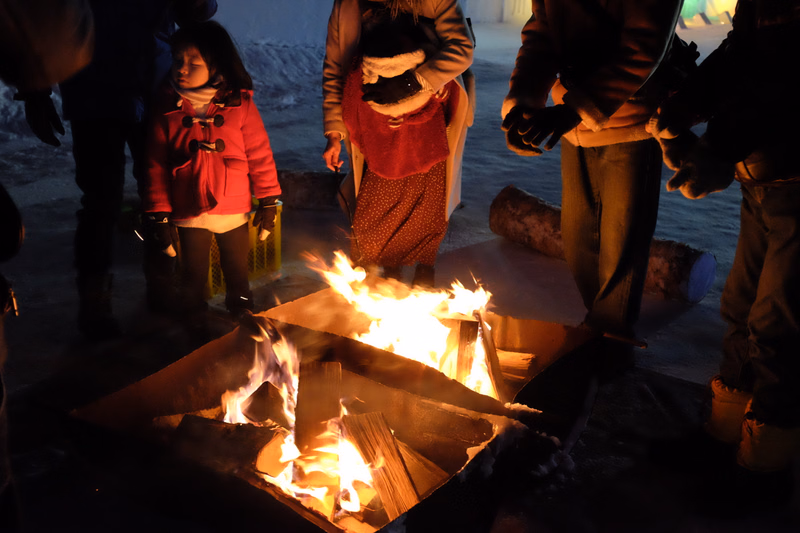 A group of people gathers around a bright campfire in the snow at night.