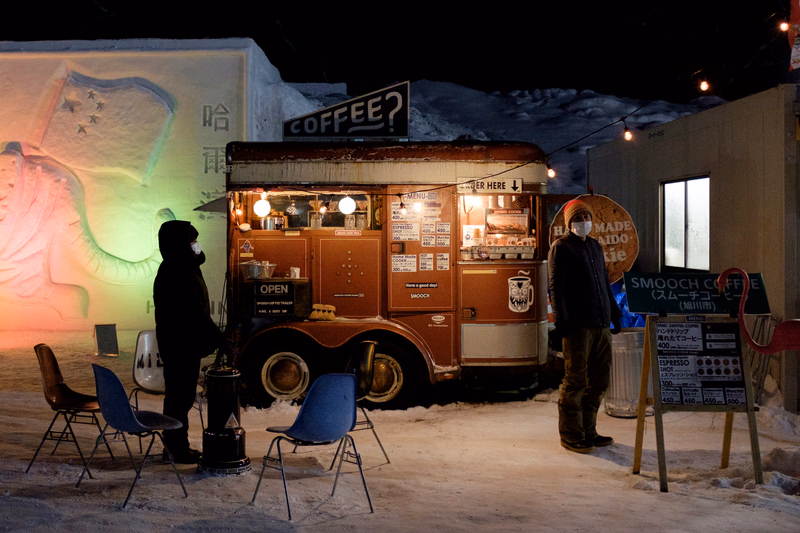 A vintage coffee cart is set up outdoors in the snow at night, with a sign that reads "COFFEE?"