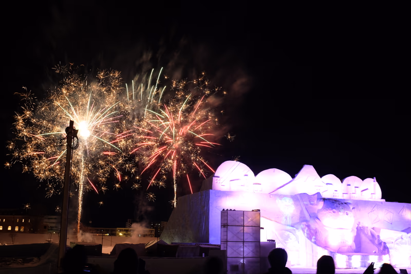 A nighttime celebration with fireworks lighting up the sky over Asahikawa, Japan. The scene is illuminated by vibrant fireworks, creating a festive atmosphere.