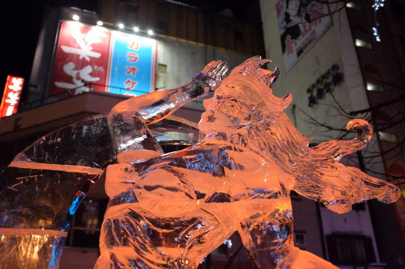 A large, detailed ice sculpture of a lion stands outdoors at night in front of a building with a sign that reads "カラオケ".