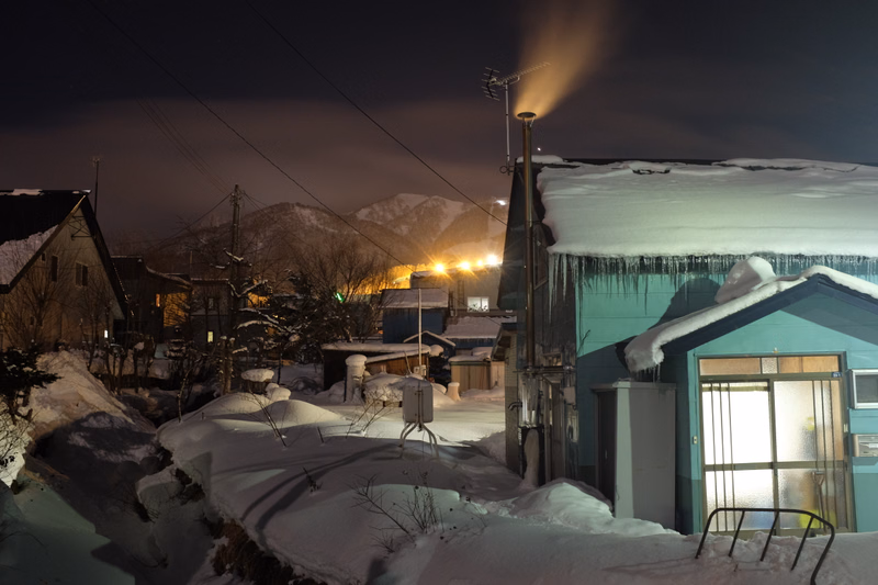 A snowy night scene shows houses with snow-covered roofs under a dark sky, with lights visible in the distance.