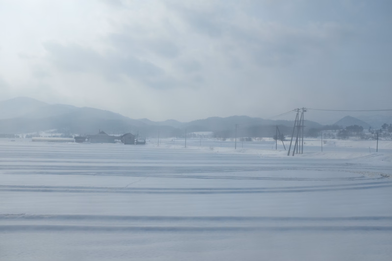 A wide, foggy winter landscape featuring a vast expanse of frozen ice under a cloudy sky, with power lines visible in the distance.