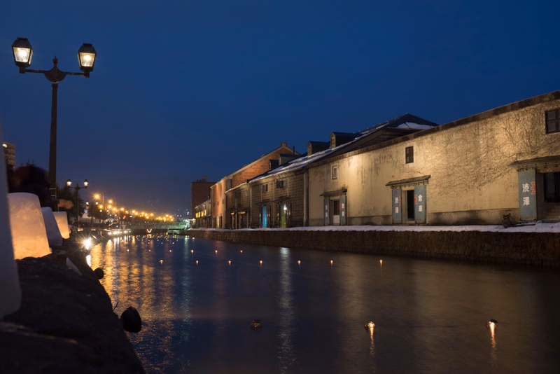 A row of traditional buildings lines a canal at night, illuminated by street lamps and small lights reflecting on the water.