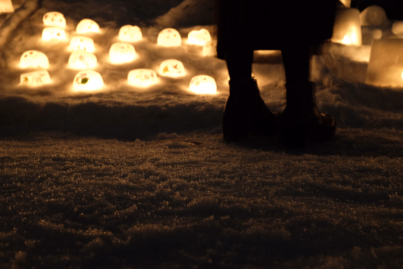 A silhouette of a person stands in the snow illuminated by a row of glowing, round lights in the background at night.