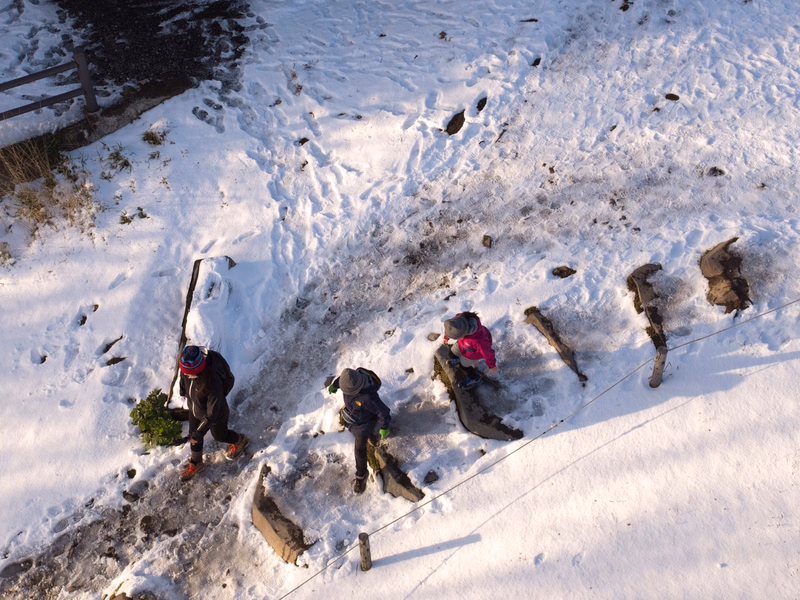 A group of hikers wearing winter gear are traversing a snowy, uneven mountain path.