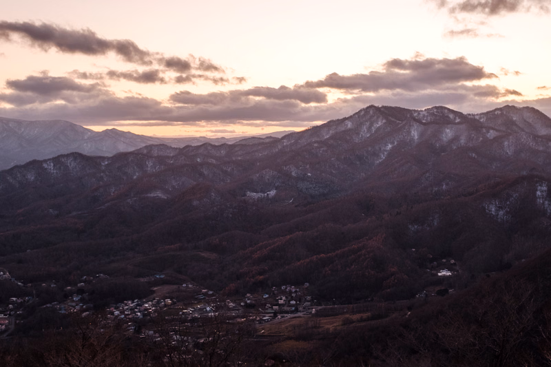 A view of rolling, dark mountains and hills under a cloudy sunset sky, with a small town visible in the valley below.