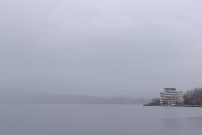 A large building is visible on the shore of a lake under a heavy, foggy sky with distant mountains.