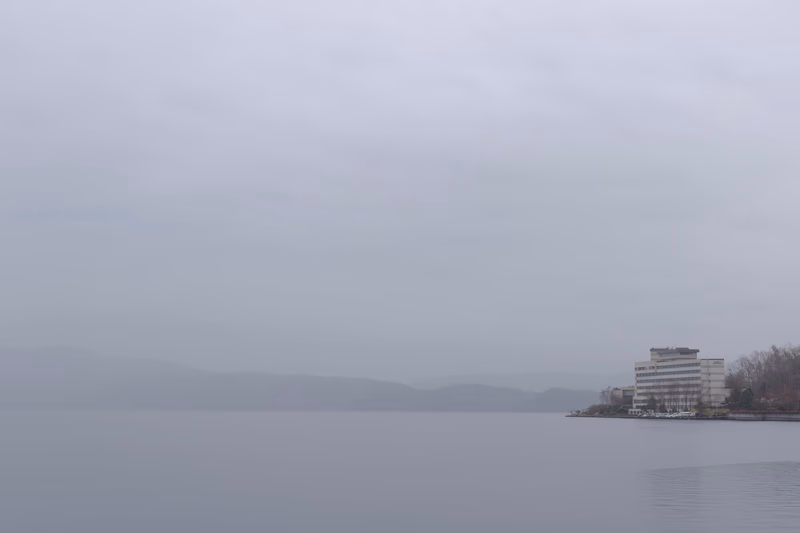 A serene landscape featuring a body of water with a distant mountain range shrouded in mist. On the right side of the image, there is a modern building with a flat roof, surrounded by trees.