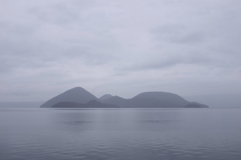 A hazy view of several mountains rising from the calm ocean under a thick, overcast sky.