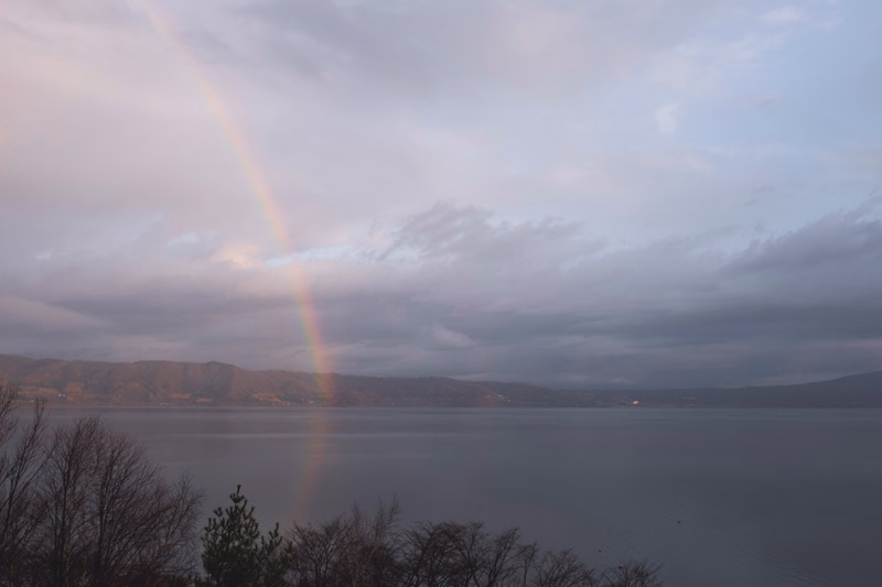 A faint rainbow arches over a large body of water under a cloudy, overcast sky.