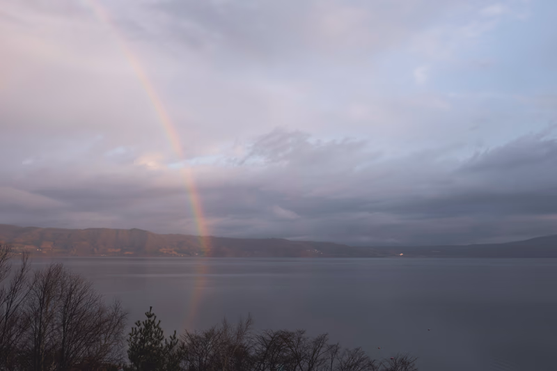 A serene lake with a rainbow arching across the sky, surrounded by mountains and trees.