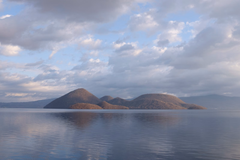 A row of rolling, tree-covered mountains sits across a calm body of water under a cloudy sky.