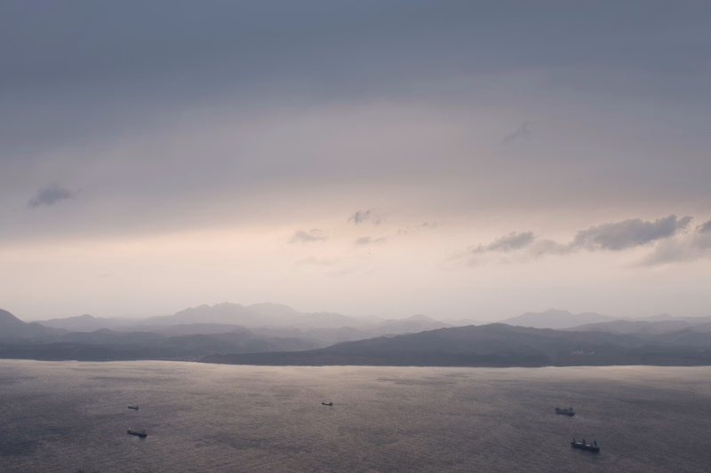 Several small ships are visible on the calm ocean under a hazy, overcast sky with distant mountains.