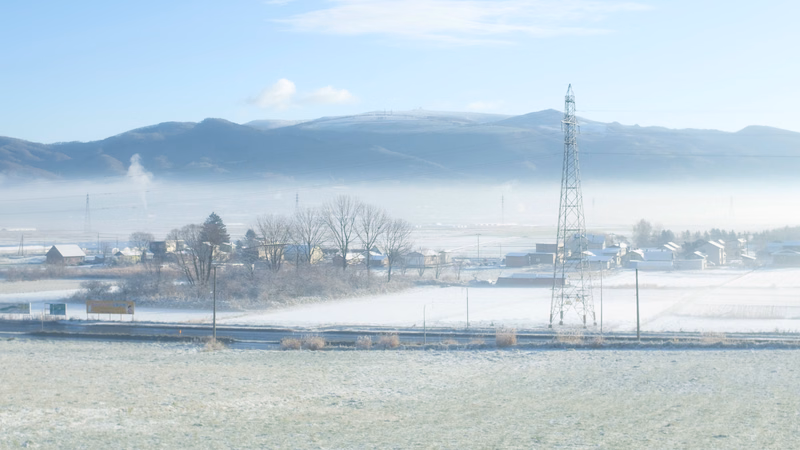 A wide view of a rural landscape with fields covered in frost, hazy mountains in the background, and several tall power lines.