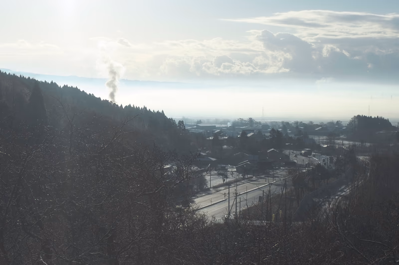 A serene winter landscape with a train passing through a village, with a plume of smoke rising from a factory in the distance.