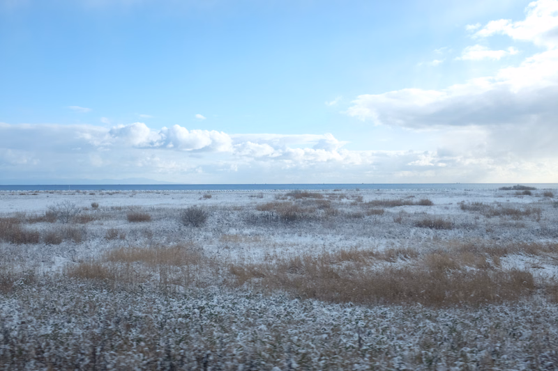 A wide, flat tundra landscape covered in patchy snow under a bright blue sky with scattered white clouds.