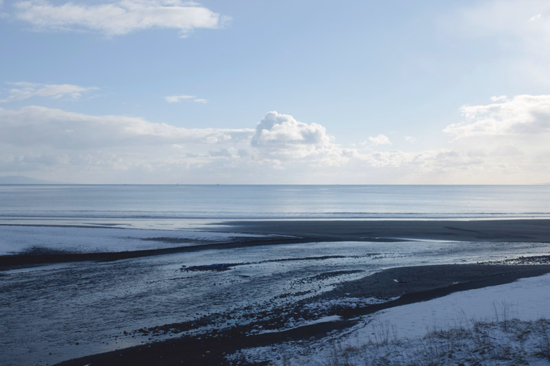 A wide, flat expanse of beach meets the calm ocean under a bright, partly cloudy sky.