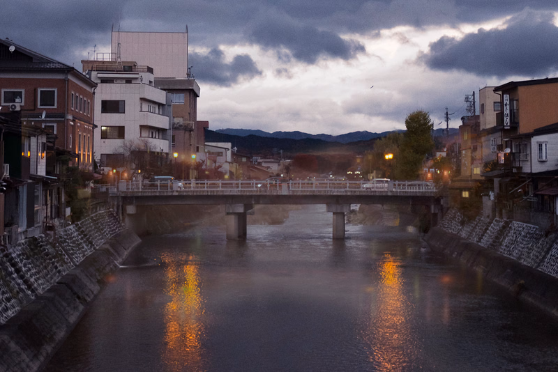 A bridge crosses a wide river in a city setting under a dramatic, cloudy sky, with buildings lining both banks.