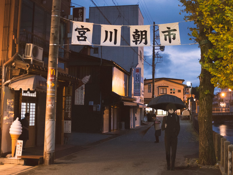 A street scene in Miyako City at dusk, featuring large banners that read "宮川市" and a person walking under an umbrella.