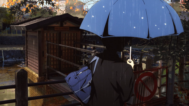 A person holding a blue umbrella stands near a wooden hut and a metal fence in the rain.