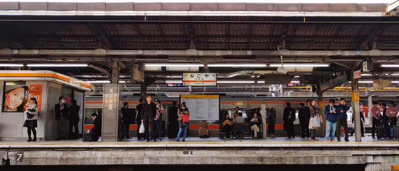 A busy train station with people waiting for a train. The platform is covered by a roof, and there are advertisements and signs visible. The people are dressed in various styles, and the atmosphere appears to be a mix of routine and anticipation.