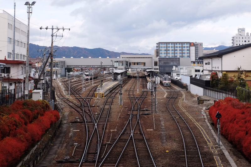A view of multiple train tracks converging in a railway station area with several buildings in the background under an overcast sky.
