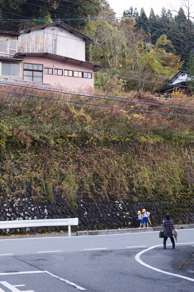 A photo of a house on a street with people walking and a tree-covered hill in the background.