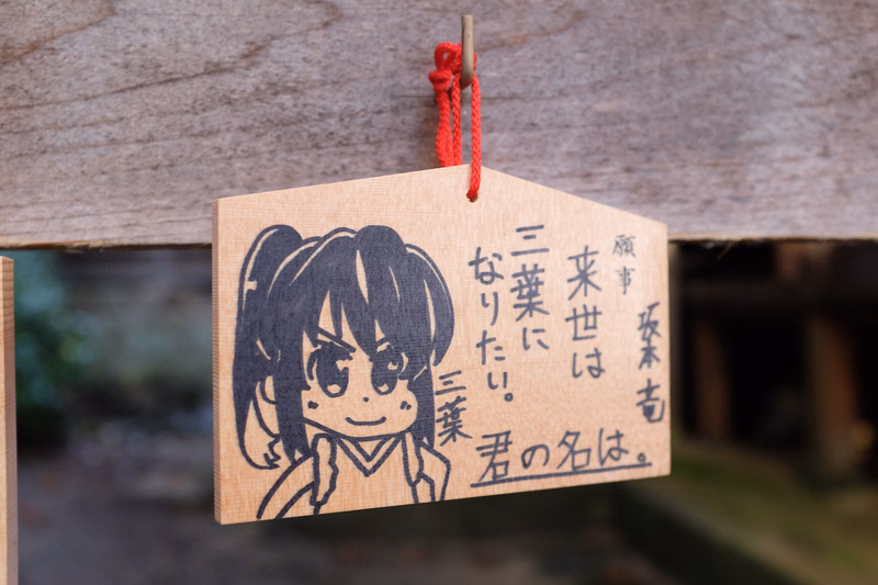 A wooden sign featuring a drawing of an anime girl and Japanese text that reads "名前は。三葉になりたい。"