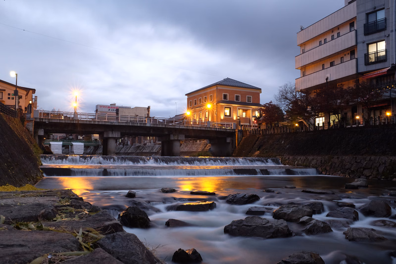 A nighttime scene featuring a low bridge over a river with a small waterfall, flanked by illuminated buildings.
