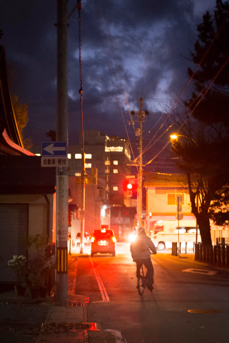 A cyclist rides down a city street at night under a dark, cloudy sky, with traffic lights visible.