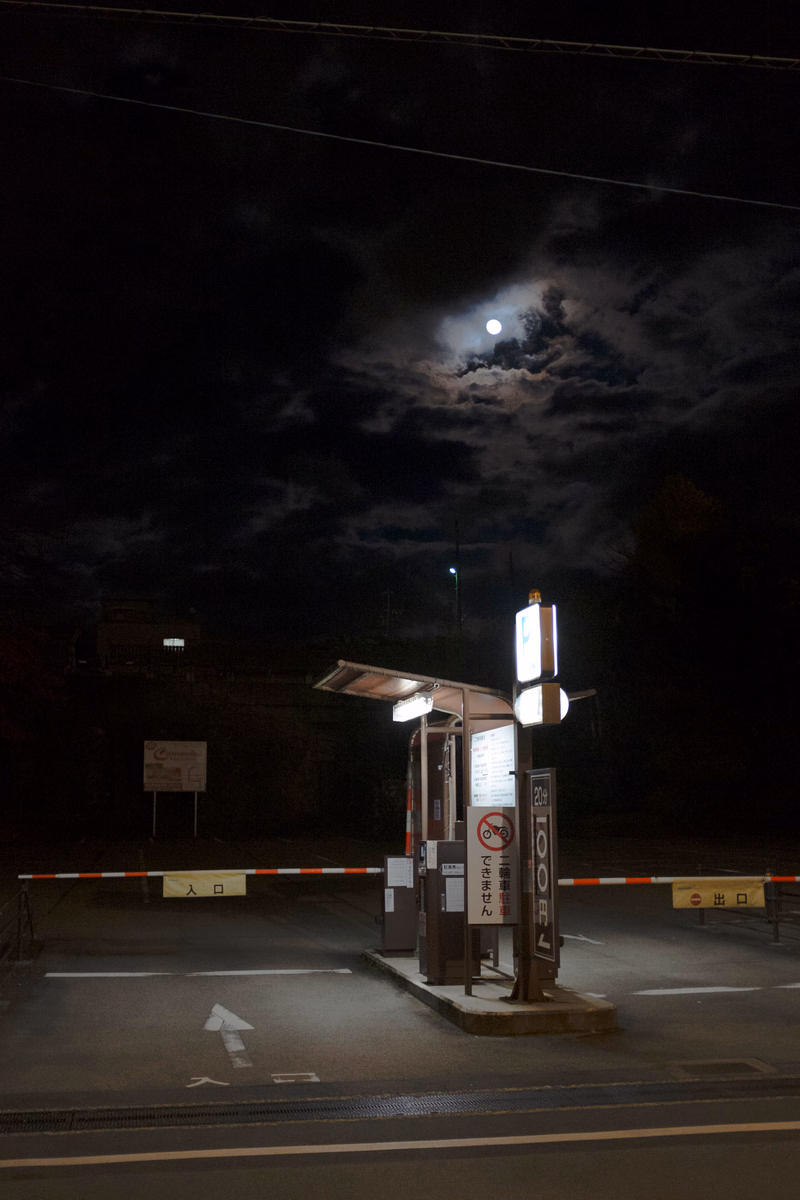 A bright moon shines through dark clouds over a nighttime entrance gate with signs reading "入口" and "出口".