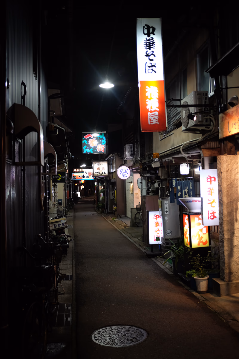 A nighttime street scene in Japan, featuring a restaurant sign and a potted plant.