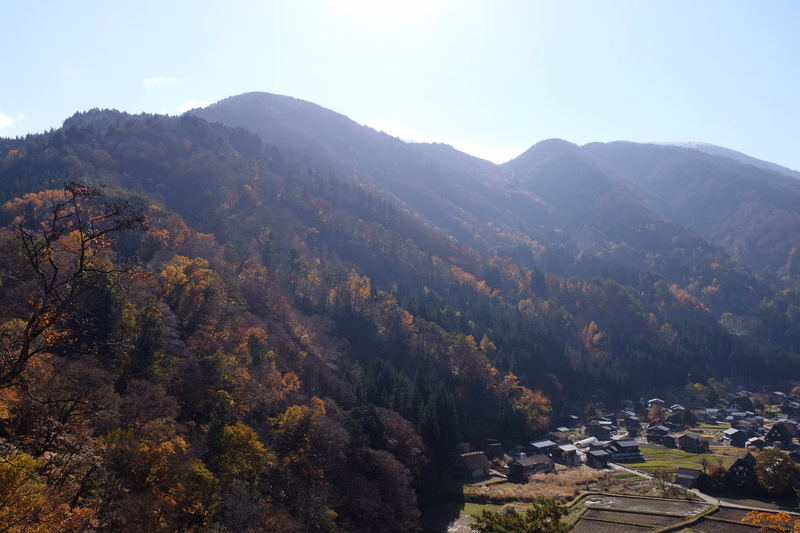 Autumn foliage covers rolling mountains and hillsides overlooking a small rural valley settlement under a bright sky.