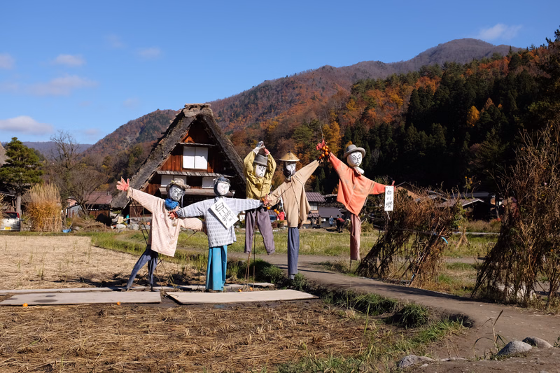 A display of various traditional festival masks stands in front of a rustic wooden hut against a backdrop of forested mountains.
