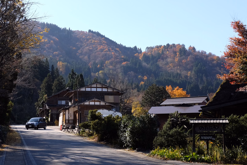A winding road passes by traditional wooden houses nestled against a backdrop of colorful autumn-covered mountains.
