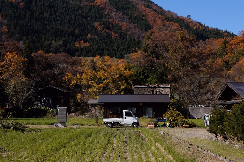 A white pickup truck is parked in front of a dark farmhouse with surrounding trees showing autumn colors against a steep mountain backdrop.