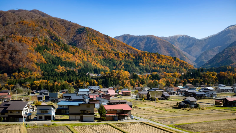 A small town nestled in a valley surrounded by mountains covered in vibrant autumn foliage under a clear blue sky.