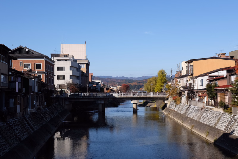 A wide river flows under a low bridge in a town lined with residential buildings under a clear blue sky.