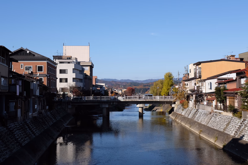 A serene view of a river in a small town with a bridge and buildings in the background, with mountains in the distance.