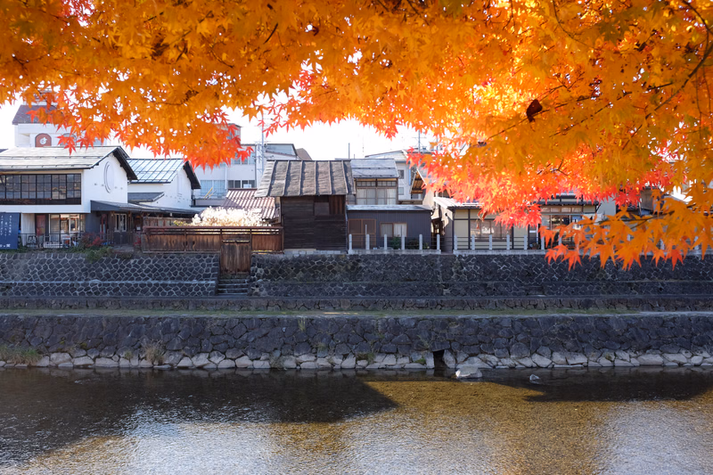Vibrant orange and red autumn leaves frame a view of traditional Japanese houses along a stone-walled riverbank.