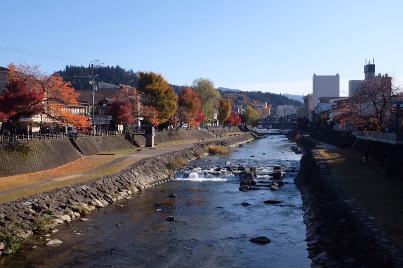 A river flows through a town lined with trees showing autumn colors under a clear blue sky.