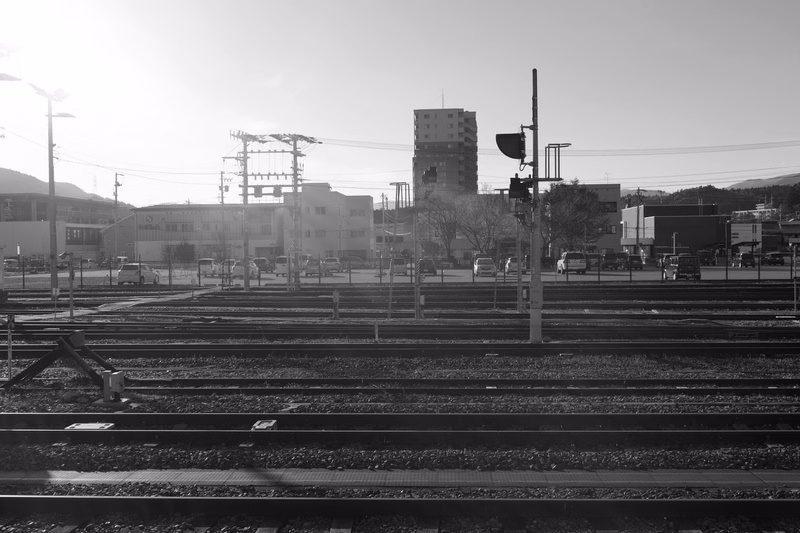 A wide view of multiple railroad tracks in an urban area, featuring overhead wires and a railway signal light.