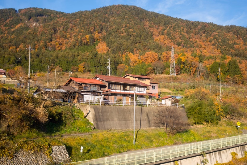 A small house is nestled against a steep, forested mountain slope during the autumn season, with train tracks visible in the foreground.