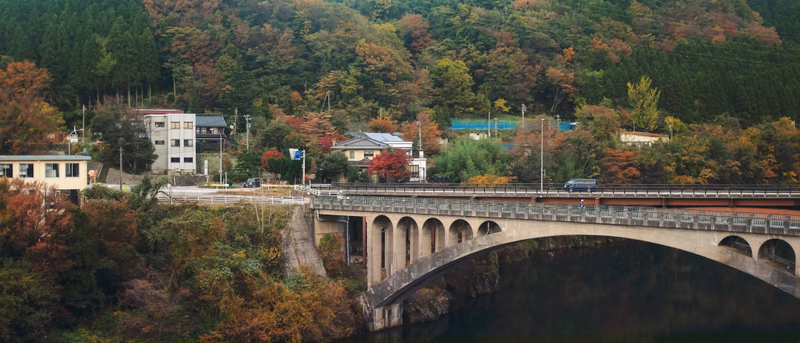 A stone arch bridge crosses a river beneath dense autumn foliage, with houses visible on the banks.