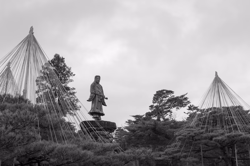 A serene garden setting with a statue and modern structures amidst traditional trees and an overcast sky.